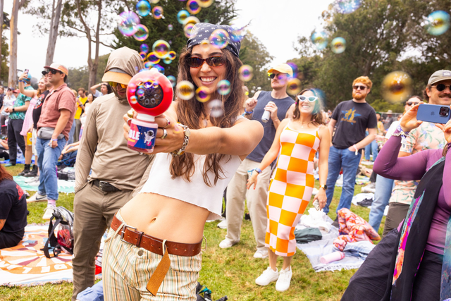 A smiling woman wearing sunglasses and a bandana blows colorful bubbles at an outdoor gathering while people in casual clothes watch and take photos.
